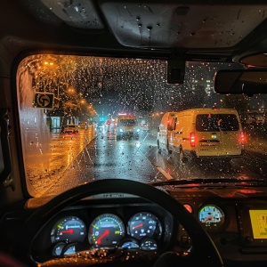 从SUV内看到的夜晚道路景象，前面是另一辆开着前灯快速行驶的面包车，可以看到雨滴落在挡风玻璃上，仪表盘上有几个仪器，包括速度计、水平指示灯，以面包车风格通过的车辆，这张照片是用iPhone Pro Max手机相机拍摄的。