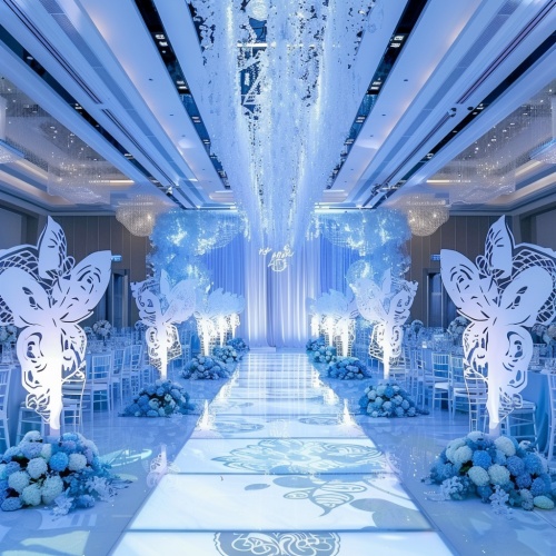 一场现代而优雅的婚礼仪式装饰,采用浅蓝色调,以白色花卉和银色饰件为特色。舞台装饰有大蝴蝶形状的装饰物,为新娘入场营造迷人的氛围。一块清晰的LED屏幕显示装饰图案,增加了整个活动空间的优雅感。在大厅的两侧,宾客坐在长桌旁,桌子用蓝色花卉布置,营造出喜庆的氛围。